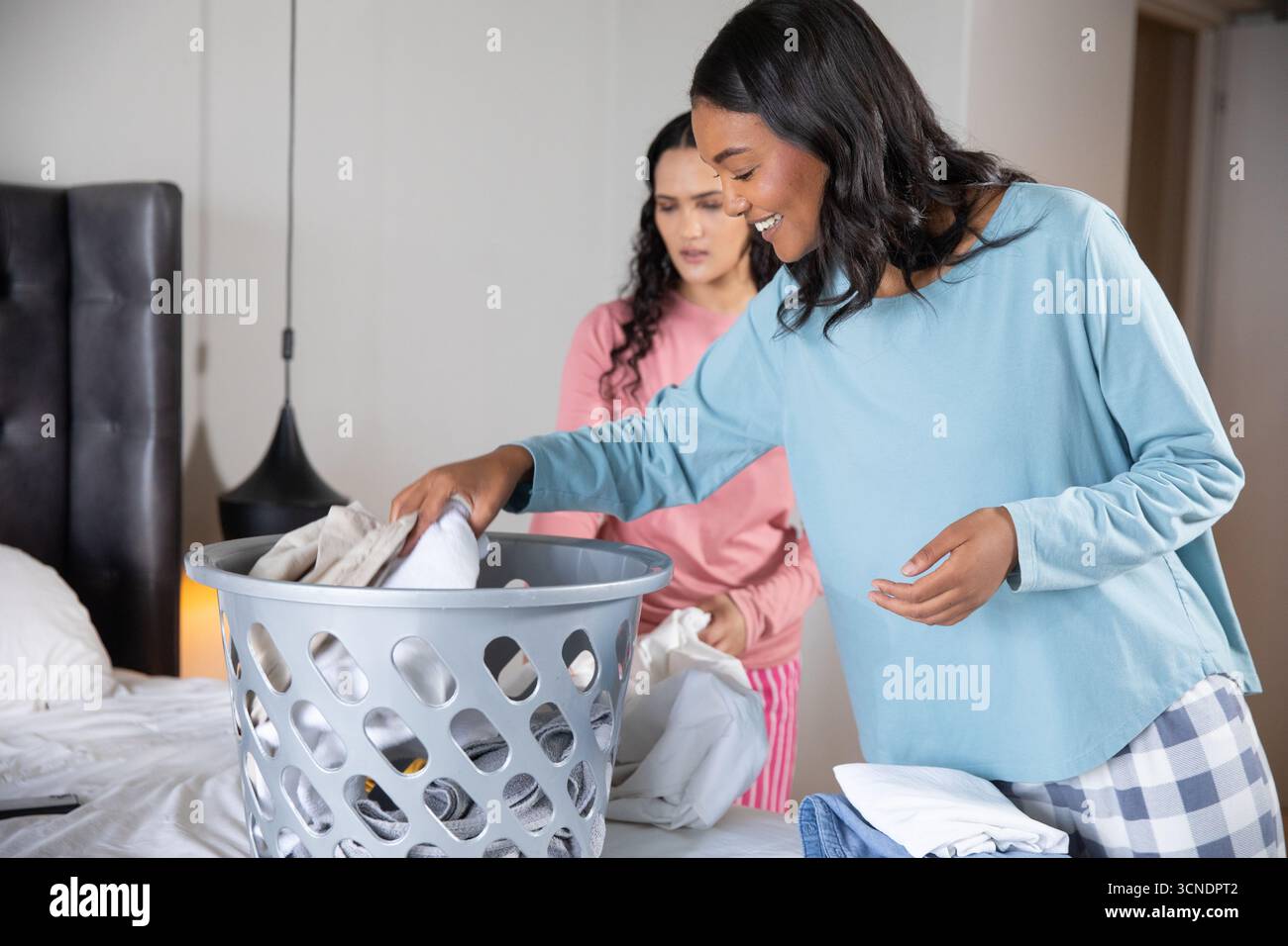Verschiedene Freundinnen im Pyjama sortieren Wäsche auf dem Bett zu Hause mit Plastikkorb Stockfoto