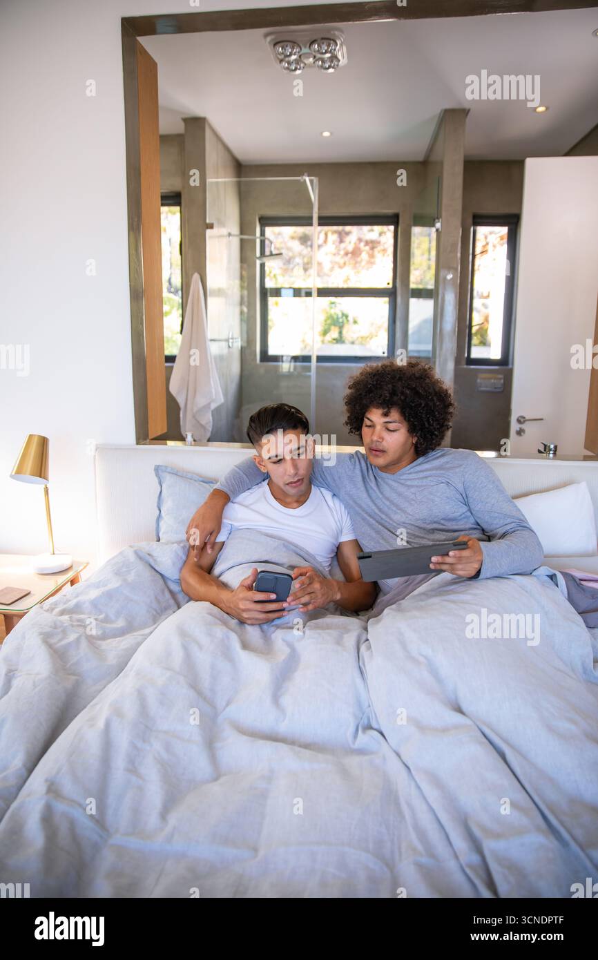 Zwei Männer sitzen unter hellgrauer Federdecke im Schlafzimmer und halten Smartphone und Tablet neben dem Beistelltisch Stockfoto