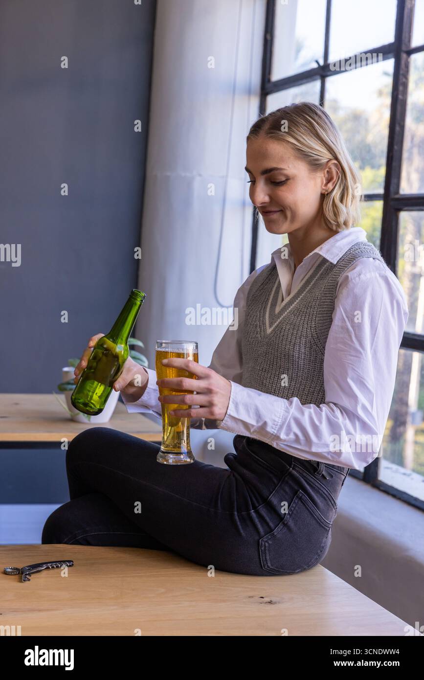 Frau, die am Tisch am Fenster sitzt und aus der Flasche in ein hohes Glas in der Nähe einer kleinen Schüssel strömt Stockfoto