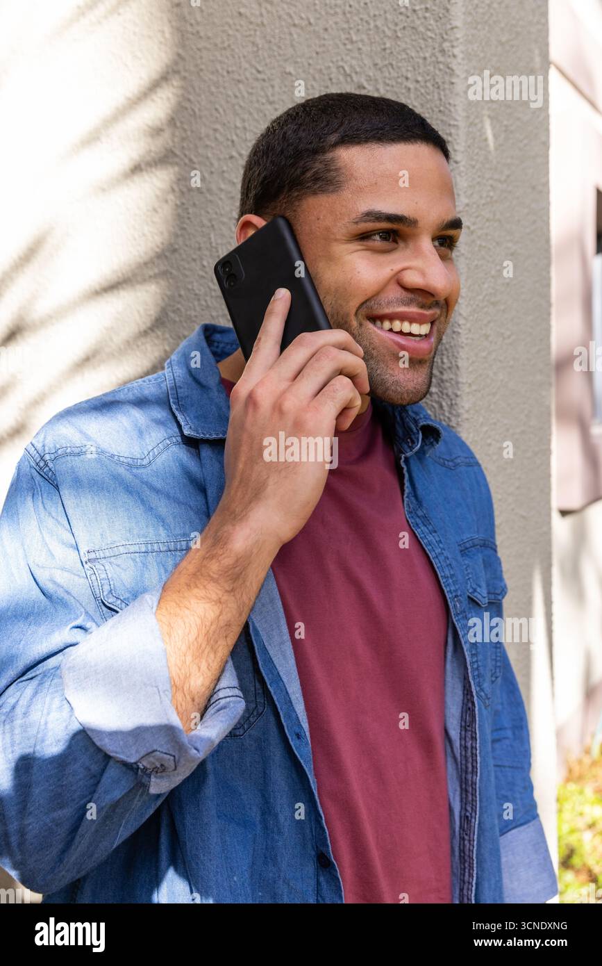 Hispanischer Mann in Denim-Hemd, der sich neben der Wand vor der Tür beugt und im Schatten der Blätter das Smartphone anruft Stockfoto