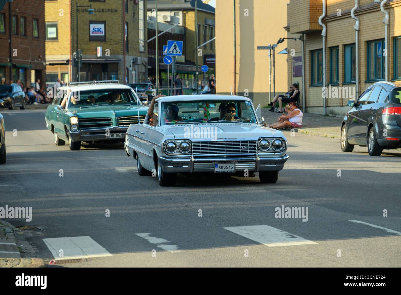 Oldtimer fahren durch eine lebhafte Stadtstraße und zeigen Vintage-Designs, während Fußgänger die Sommeratmosphäre genießen. Die Sonne scheint und schafft ein che Stockfoto