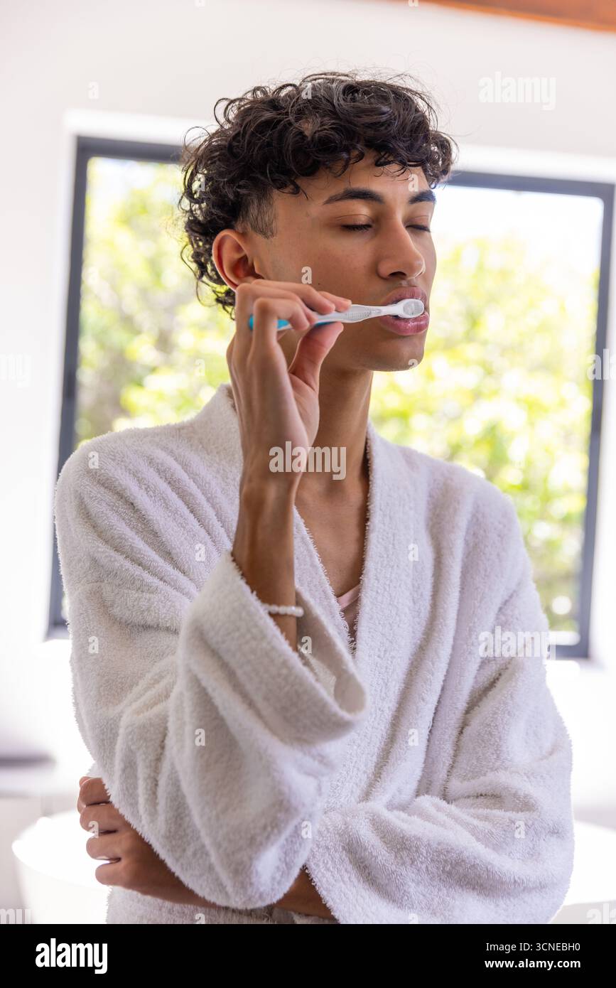 Mann, der in der Nähe des Badezimmerfensters steht, putzt Zähne mit weißer Zahnbürste und trägt einen weichen Bademantel Stockfoto