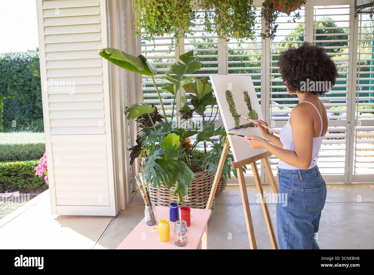 afroamerikanerin, die botanische Formen an der Staffelei auf der überdachten Veranda mit Farbgläsern malte Stockfoto