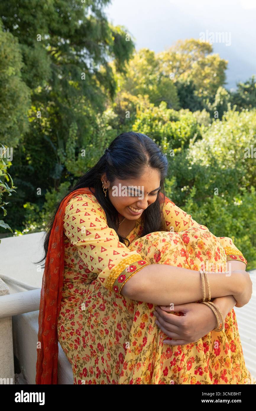 Indianerin mit gelber Kurta, rotem Schal, sitzt auf Balkonvorsprung und umarmt die Knie, lächelt Stockfoto