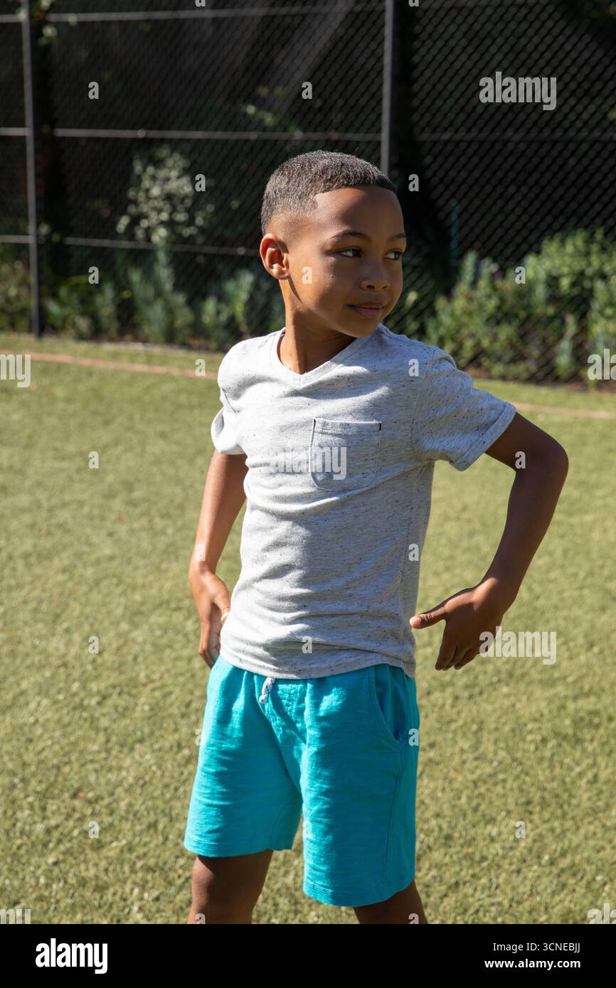 Afroamerikanischer Junge, der Sportswear auf dem Rasen in der Nähe des Maschendrahtzauns trägt, sieht nach rechts aus und ruht Hände Stockfoto