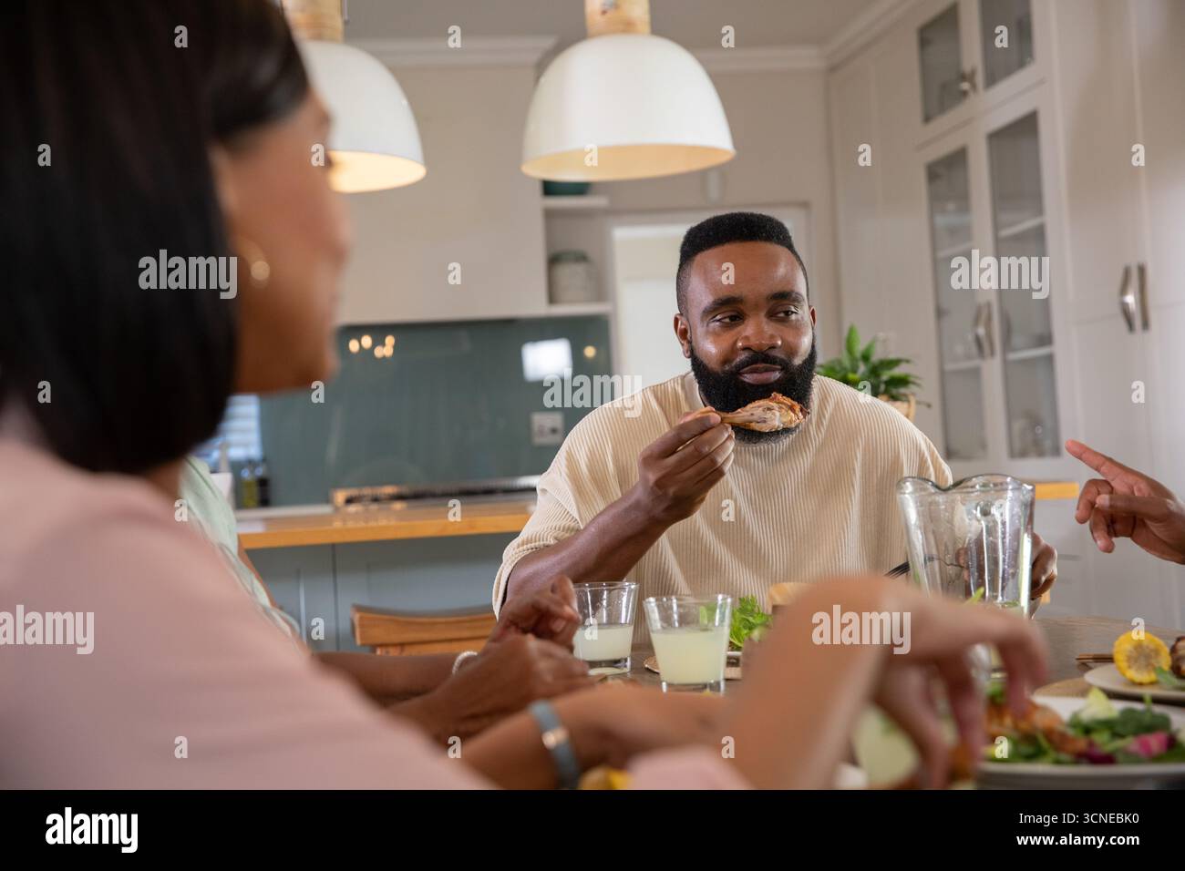 Verschiedene Freunde essen Hähnchenflügel und plaudern um einen hölzernen Esstisch in der modernen Küche Stockfoto