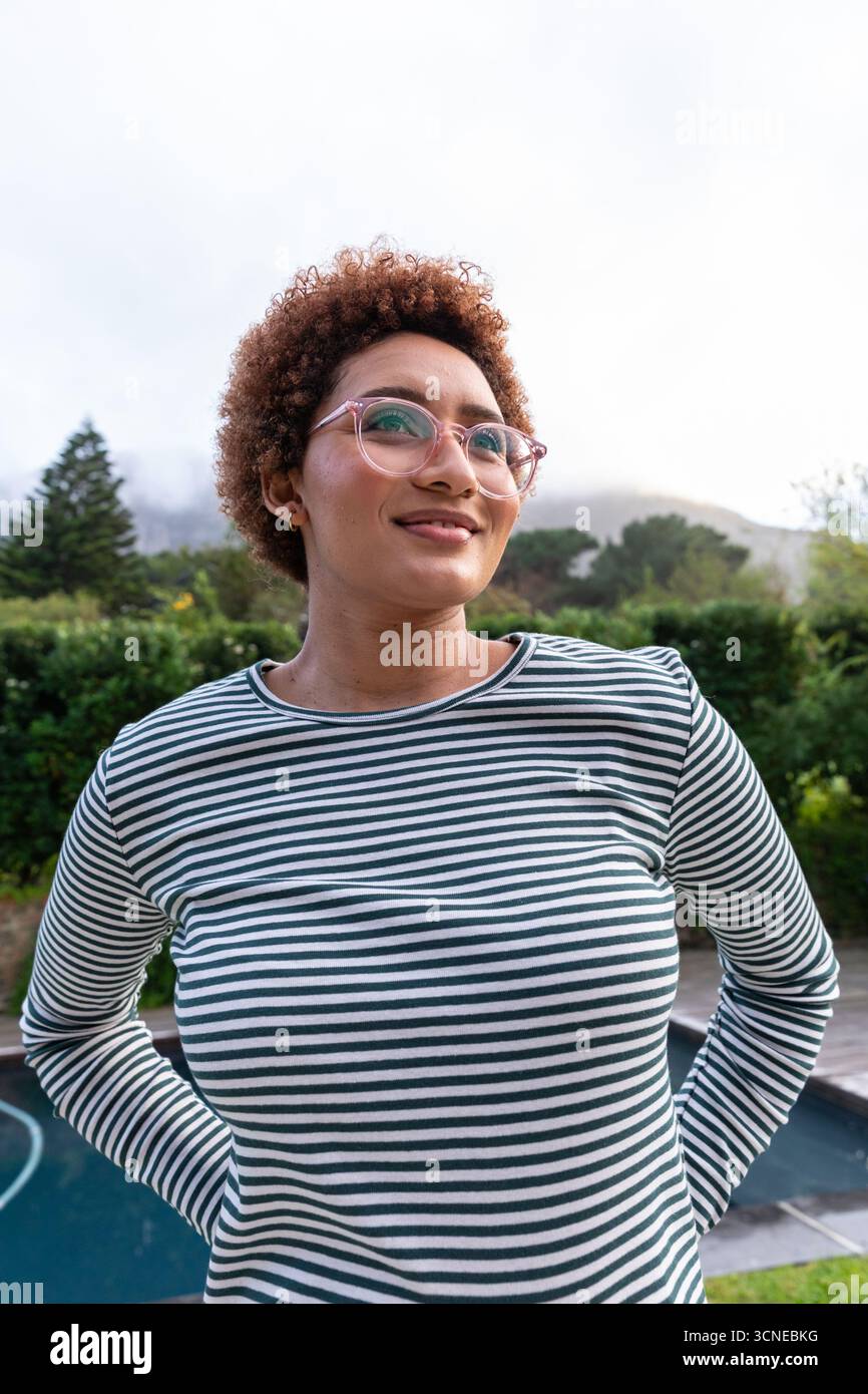 afroamerikanerin, die neben dem Pool im Garten steht und ein gestreiftes Hemd und eine Brille trägt Stockfoto