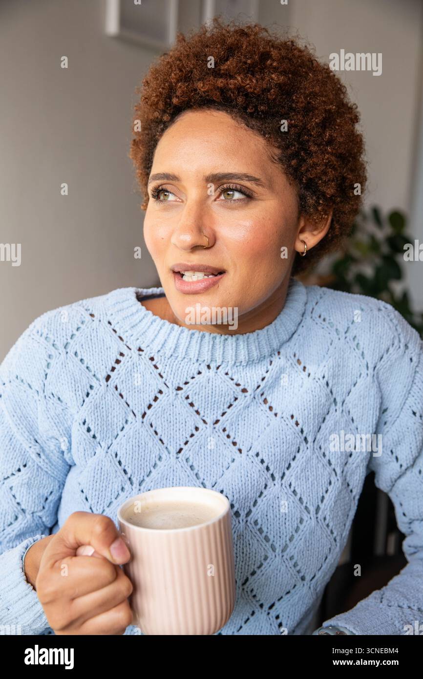 afroamerikanerin, die zu Hause einen gerippten Becher hält und links gerahmtes Kunstwerk betrachtet, Pflanze Stockfoto