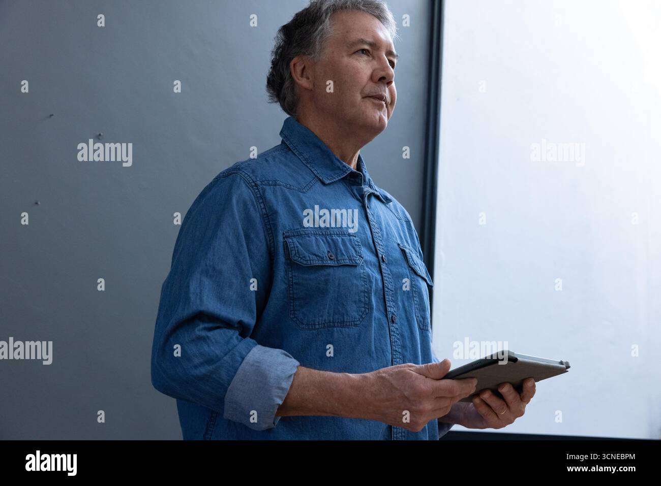 Mann, der in einem modernen Büro in einem Denim-Hemd steht und in Richtung Fenster blickt Stockfoto