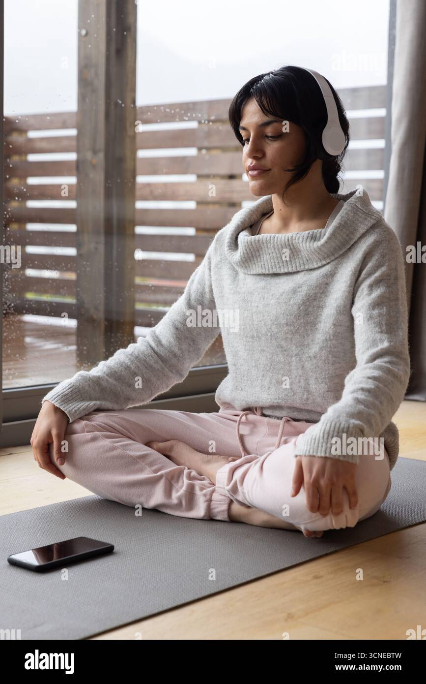 Frau übt Meditation sitzend auf Yogamatte durch Glastür mit Kopfhörern, Tablet Stockfoto