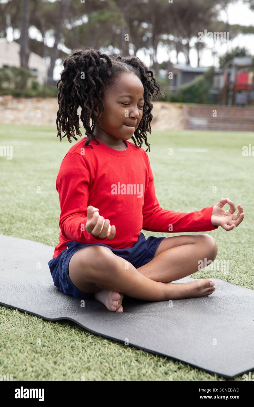 afroamerikanisches Kind in rotem Hemd und marineblauen Shorts, das auf schwarzer Matte im öffentlichen Park meditiert Stockfoto