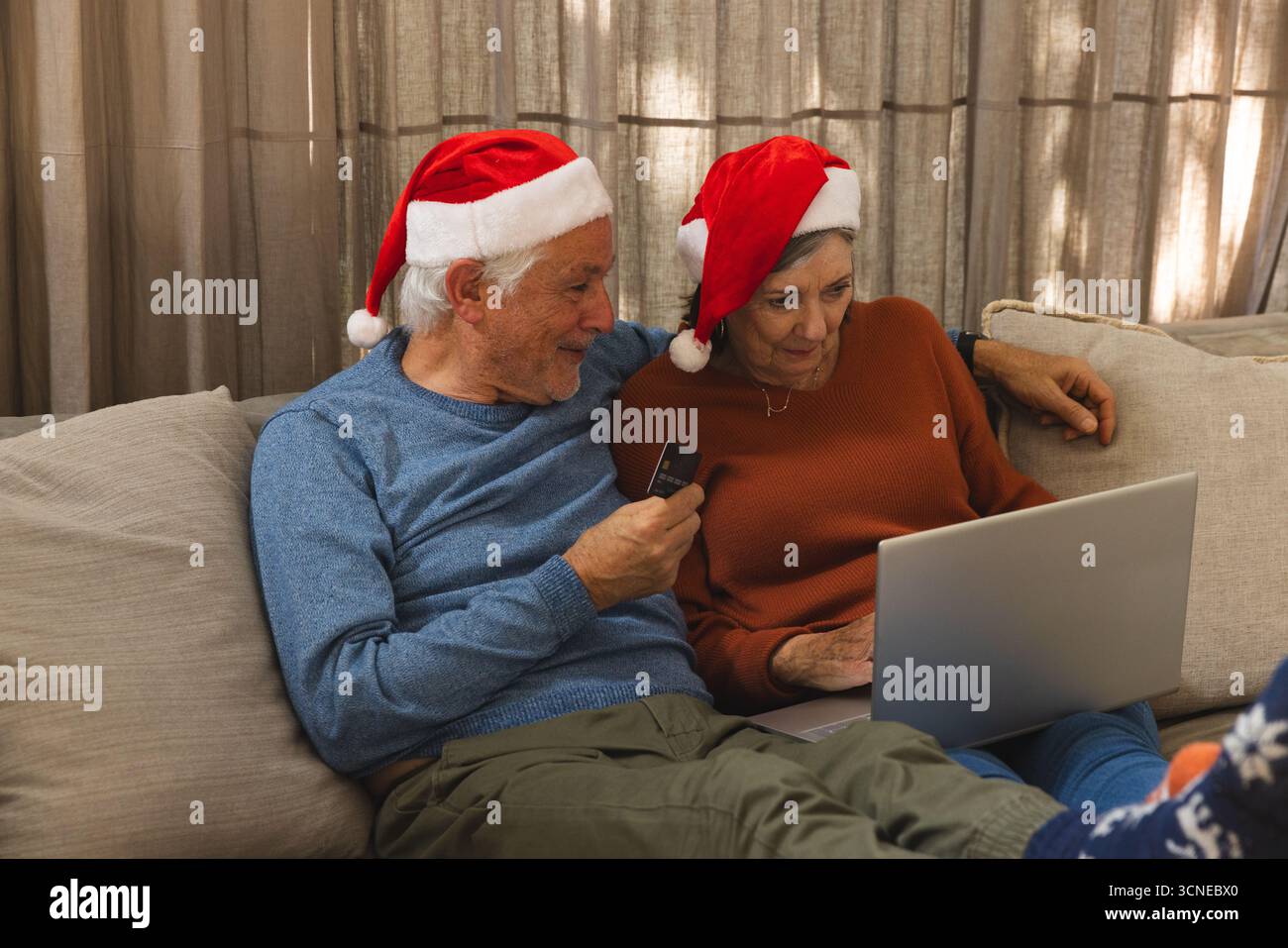 Seniorenpaar mit Weihnachtsmützen beim Online-Shopping mit silbernem Laptop und Kreditkarte auf dem Sofa Stockfoto