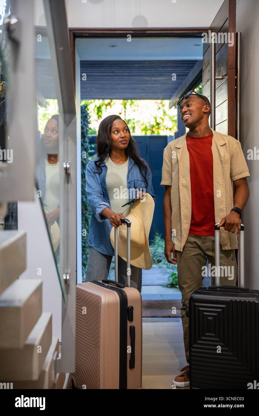 Afroamerikanisches Paar, das im Eingangsbereich steht und Koffer mit Strohhut hält und Sonnenbrille trägt Stockfoto