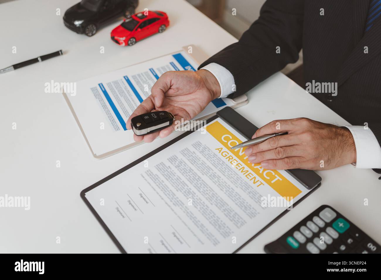 Ein Geschäftsmann in einem formellen Anzug erklärt die Details der Autoversicherung, hält ein kleines rotes Auto-Modell und zeigt auf Vertragsdokumente, was eine Einigung symbolisiert Stockfoto