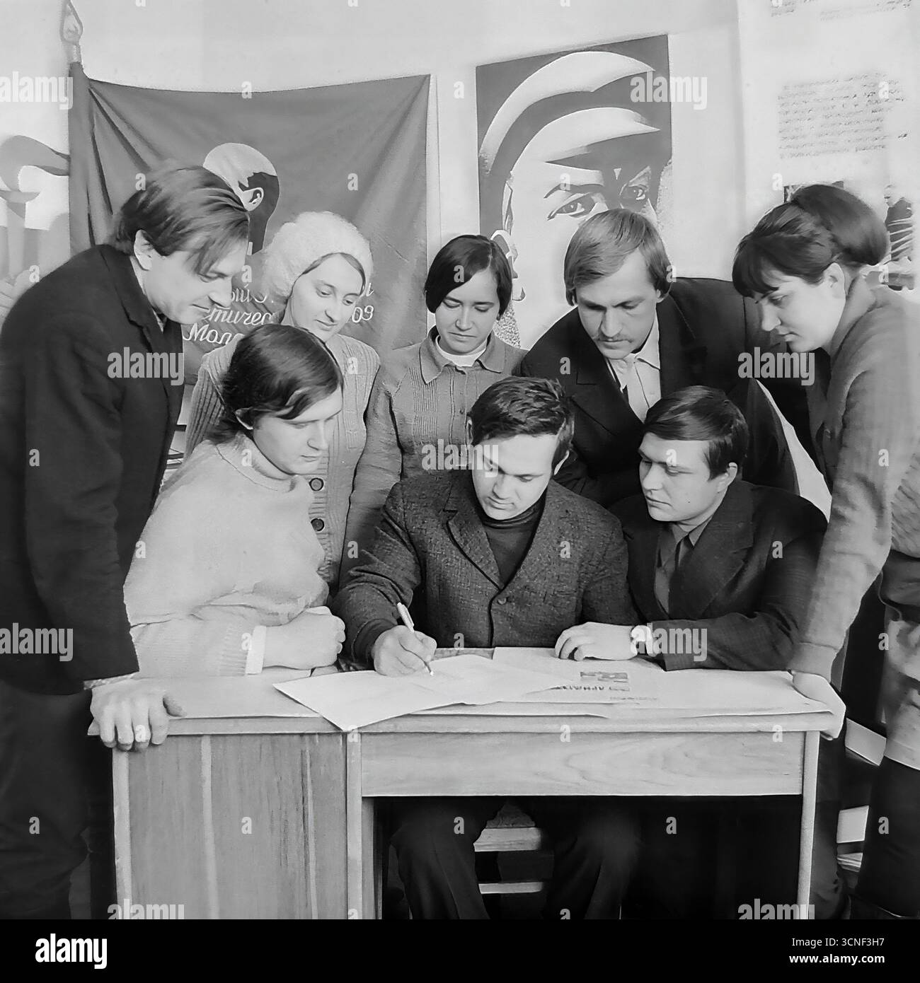 Junge Arbeiter nehmen in den 1970er Jahren an einem Treffen der Komsomol (Junge Kommunistische Liga) in der „Roten Ecke“ im AIZ-Werk in Sloviansk, UdSSR Teil Dieses Schwarzweiß-Archivfoto zeigt die Szene der Unterzeichnung eines wichtigen Dokuments vor dem Hintergrund sowjetischer Plakate und des Komsomol-Banners, das die Atmosphäre des ideologischen und sozialen Lebens der sowjetischen Jugend vermittelt Stockfoto