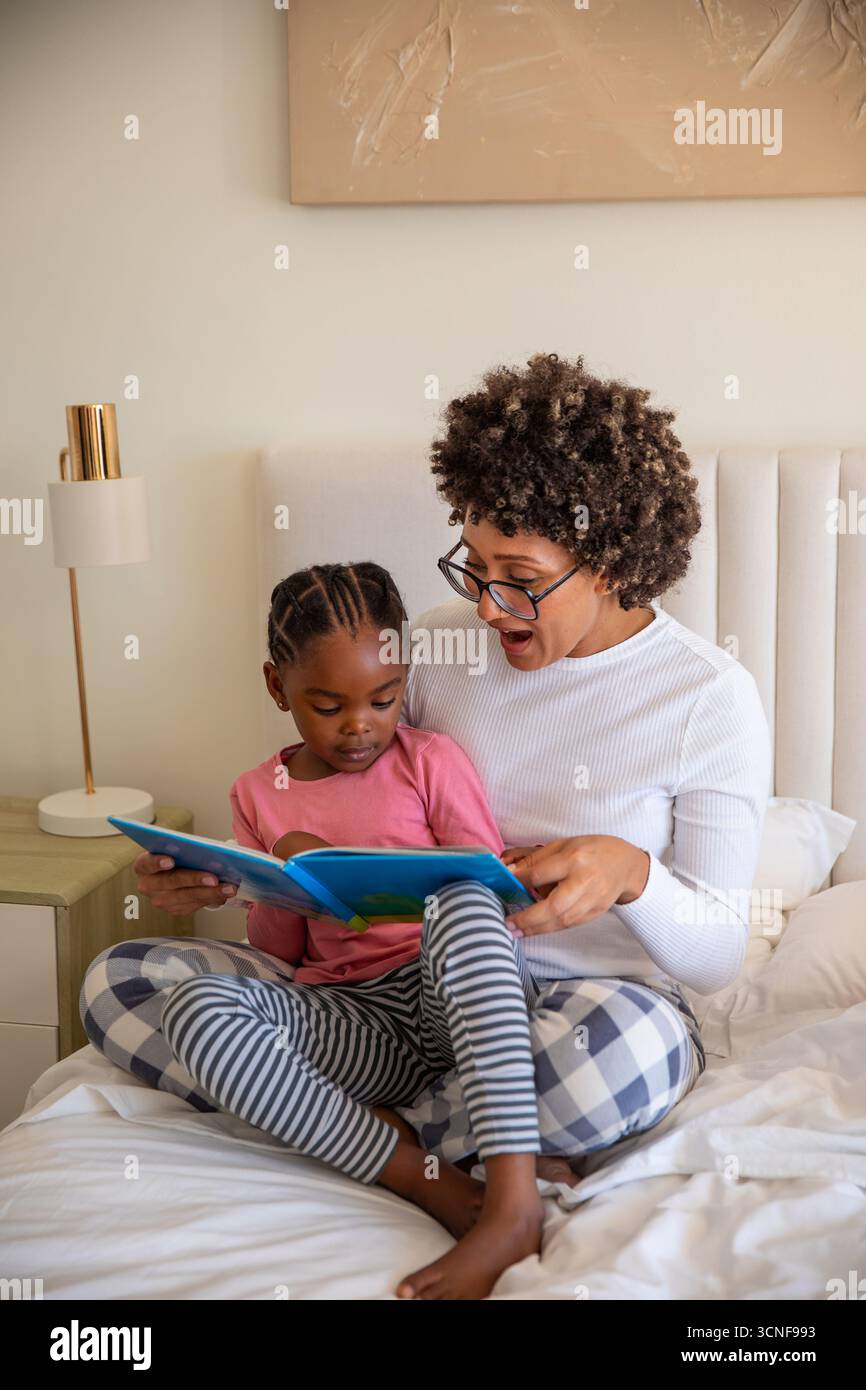 afroamerikanische Mutter und Tochter lesen Kinderbuch auf Bett mit Kopfteil und Lampe Stockfoto