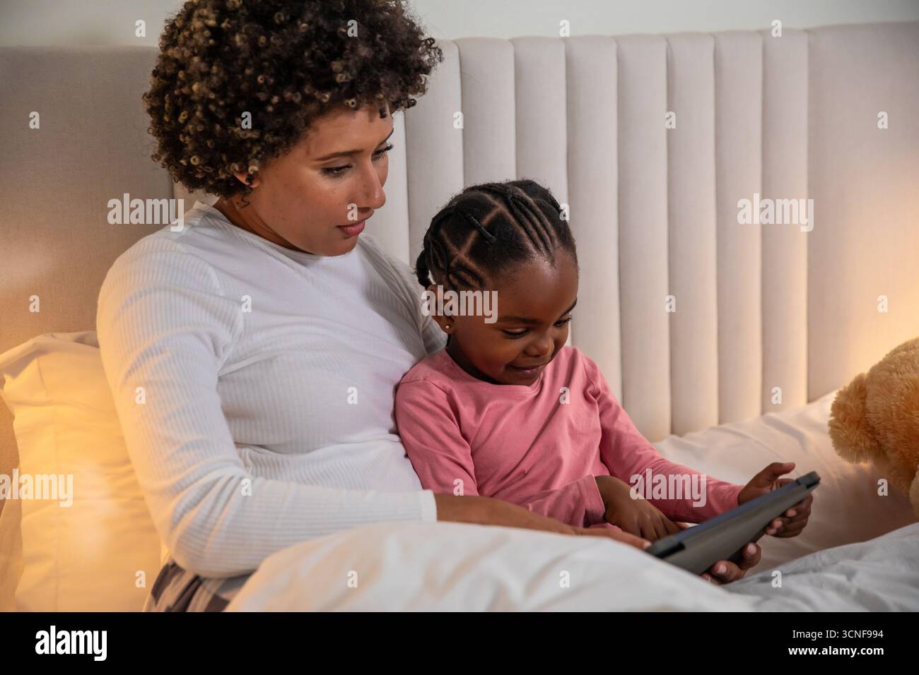 Mutter mit afroamerikanischer Tochter, die sich ein Tablet unter Nachttischlampe auf dem Bett mit Teddybär teilt Stockfoto