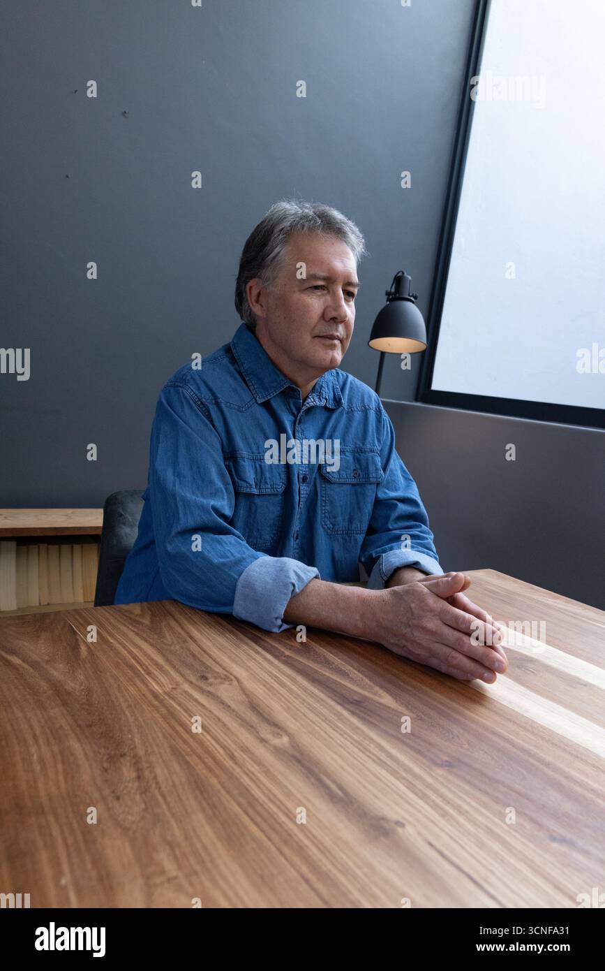 Mann sitzt am hölzernen Arbeitstisch unter einem großen Fenster und hält Hände nahe der Lampe Stockfoto