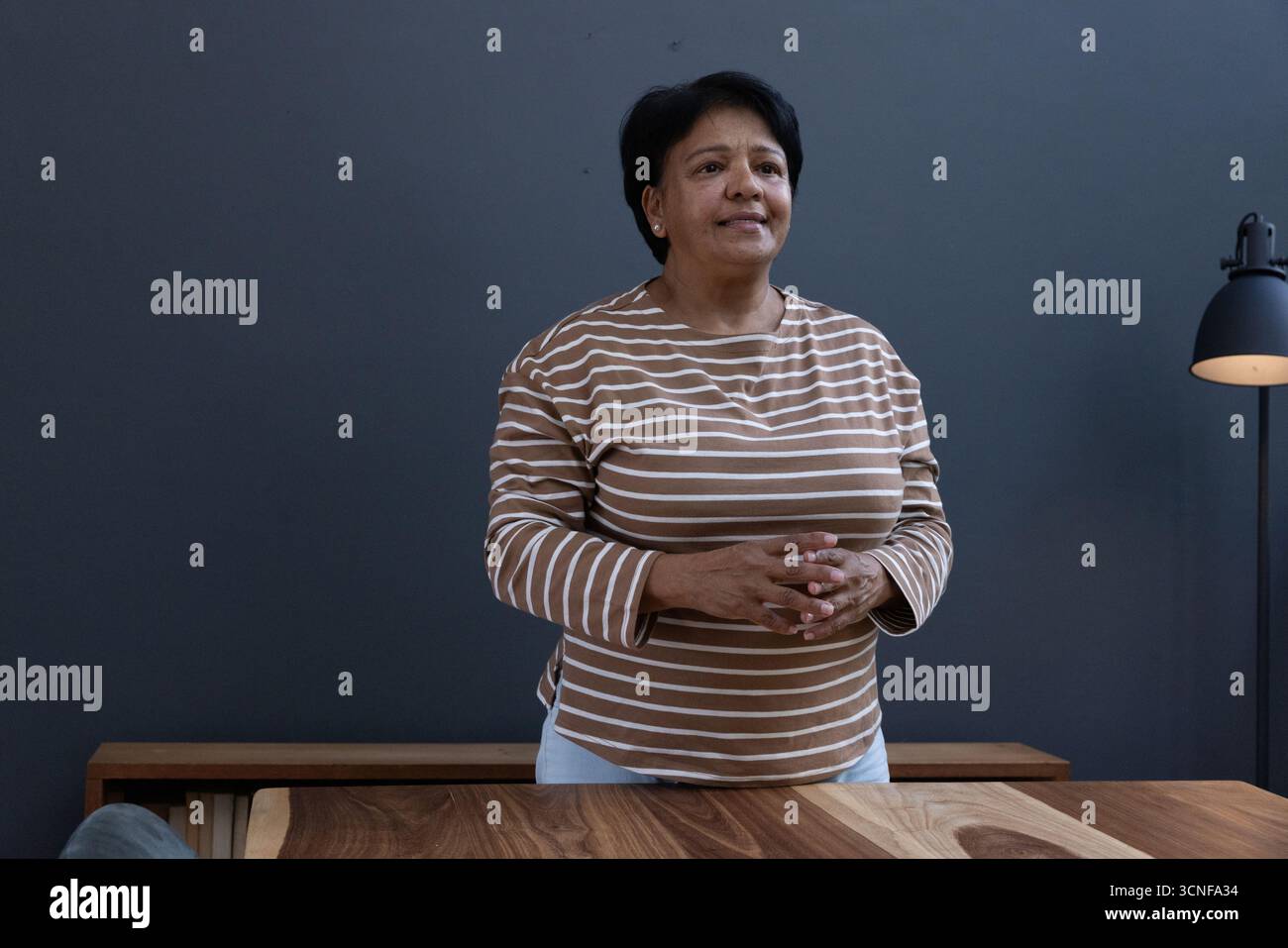 Ältere indische Frau, die hinter Holztisch mit Stehlampe vor grauer Wand zu Hause steht Stockfoto