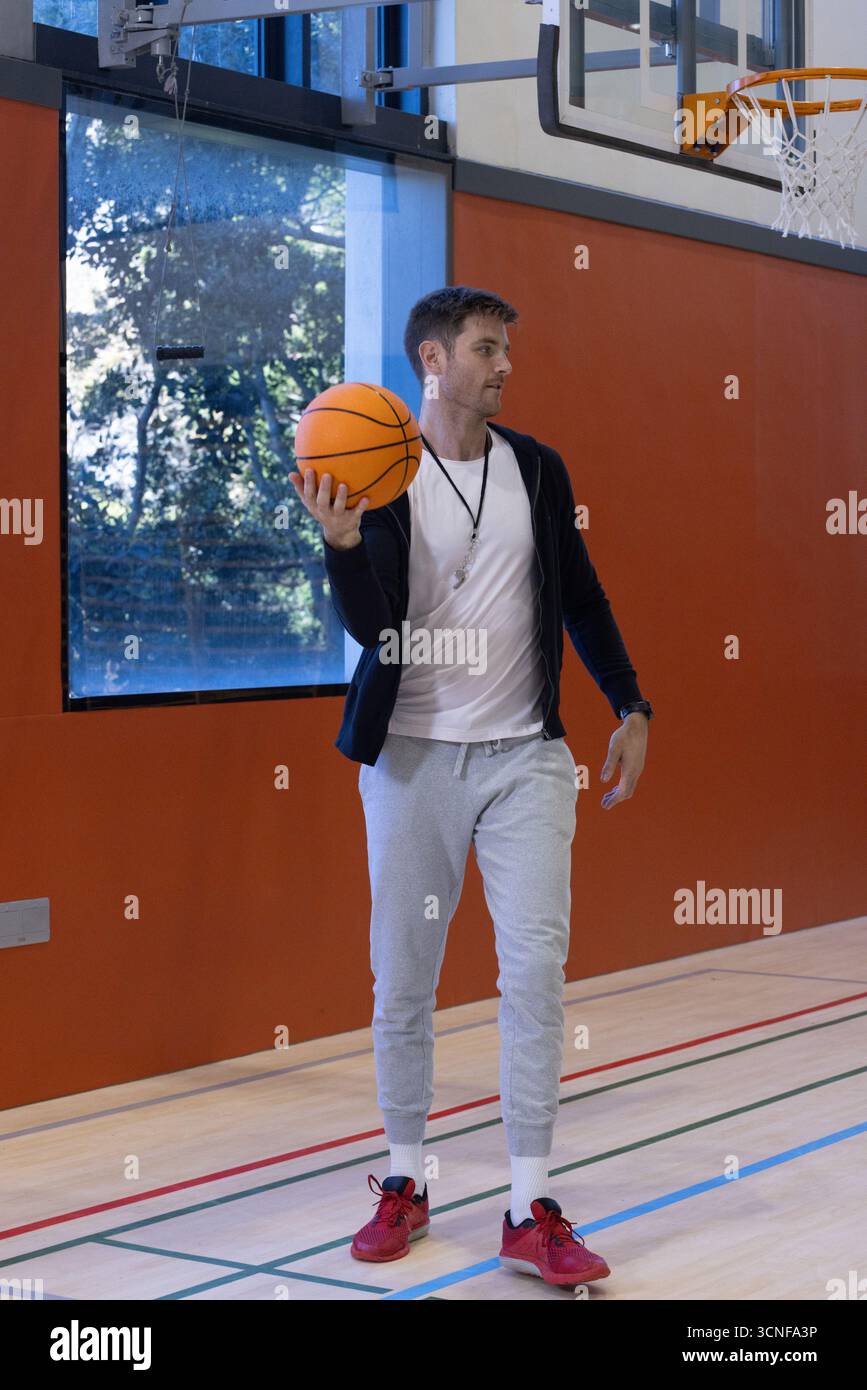 Basketballtrainer hält den Ball auf einem polierten Platz in der Nähe von Fenstern und montiertem Korb Stockfoto