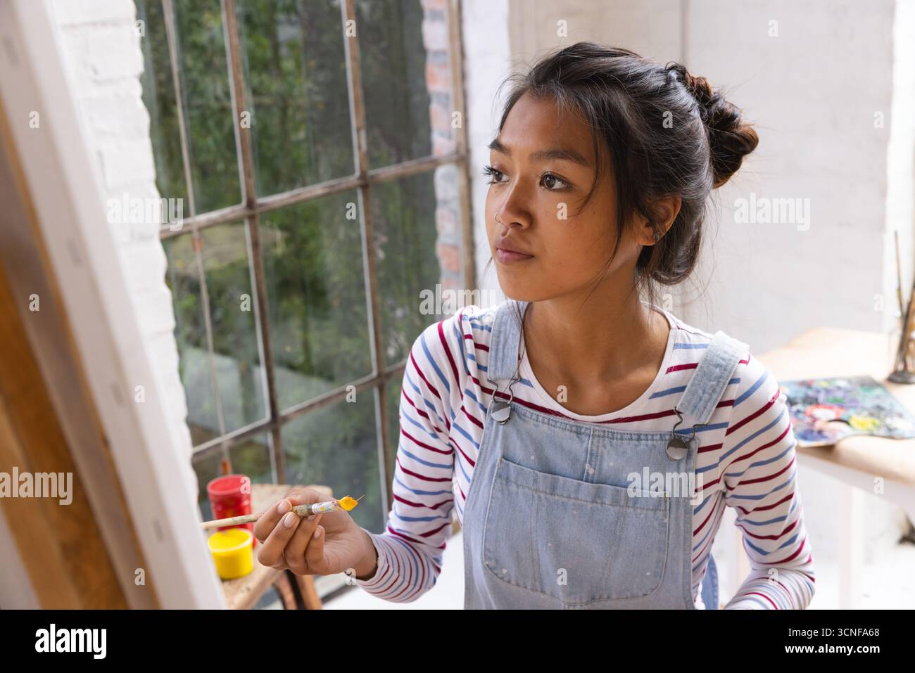 Asiatische Malerin in Overalls, die den Pinsel am Atelierfenster über dem Arbeitstisch hält, Kopierraum Stockfoto
