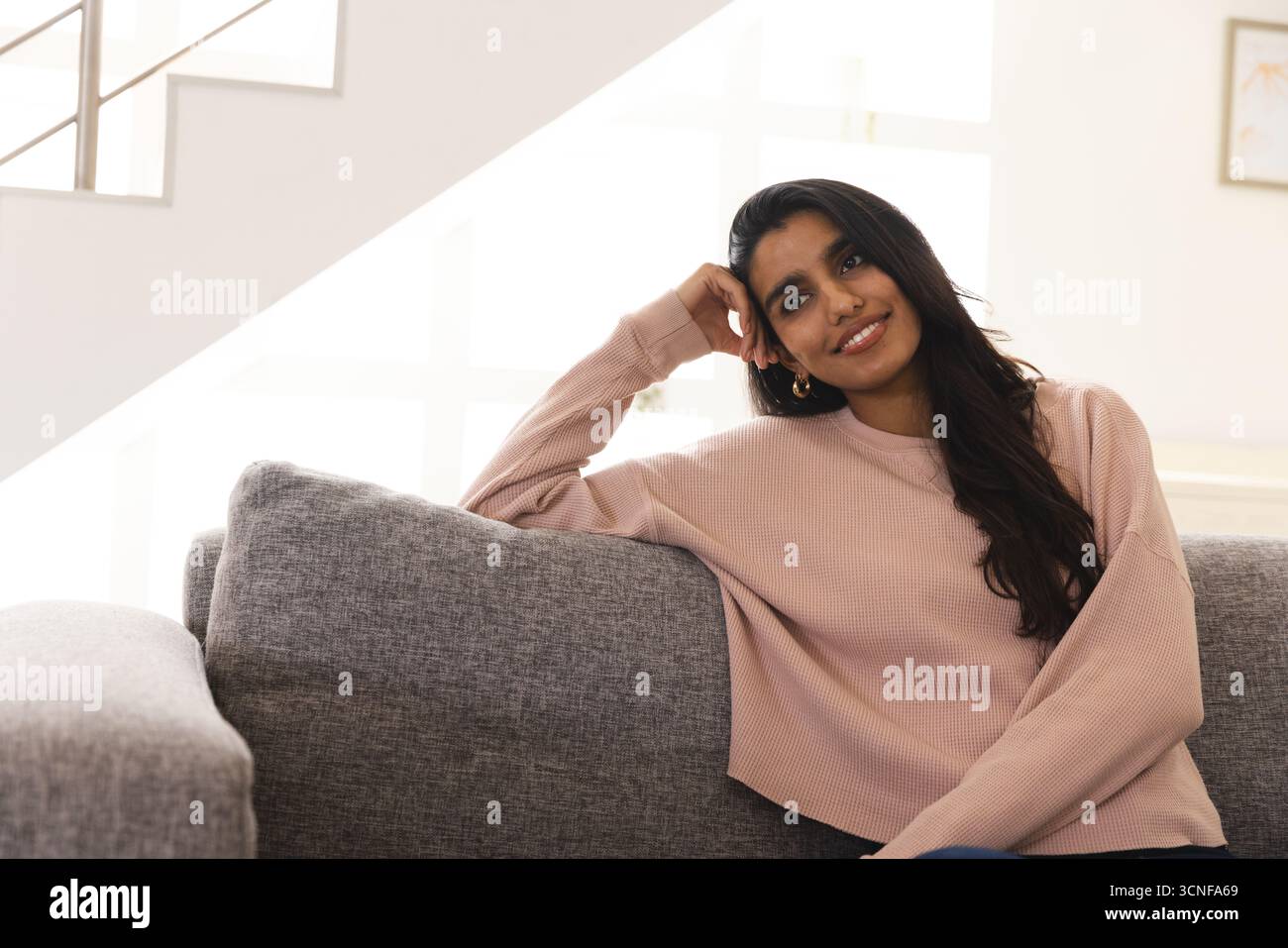 Südasiatische Frau, die sich zu Hause auf dem grauen Sofa stützt, links auf das Fenster mit mehreren Fensterscheiben blickt, Kopierraum Stockfoto