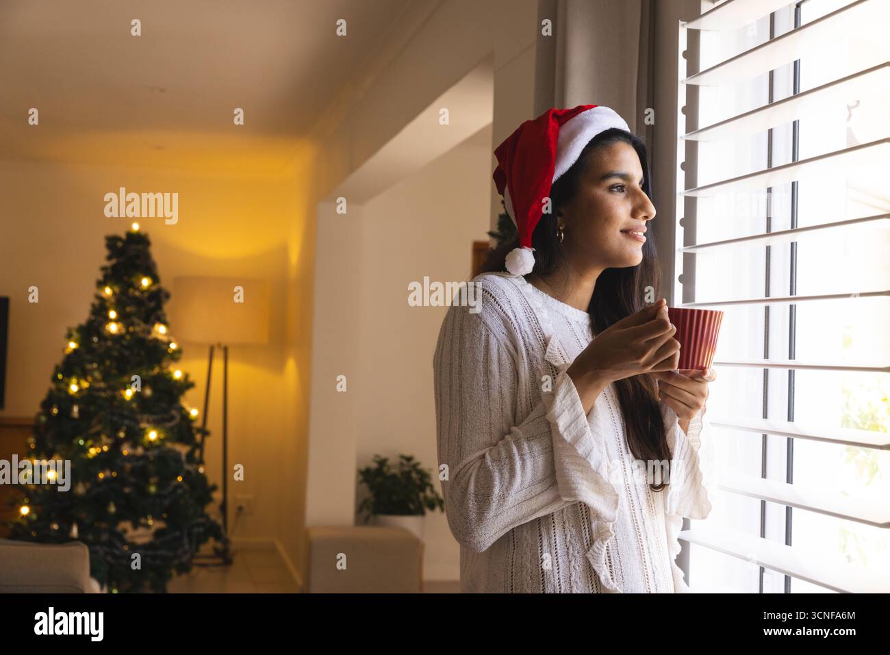 Frau mit Weihnachtsmütze, Pullover, stehend bei Jalousien im Wohnzimmer, mit rotem Becher, Kopierraum Stockfoto