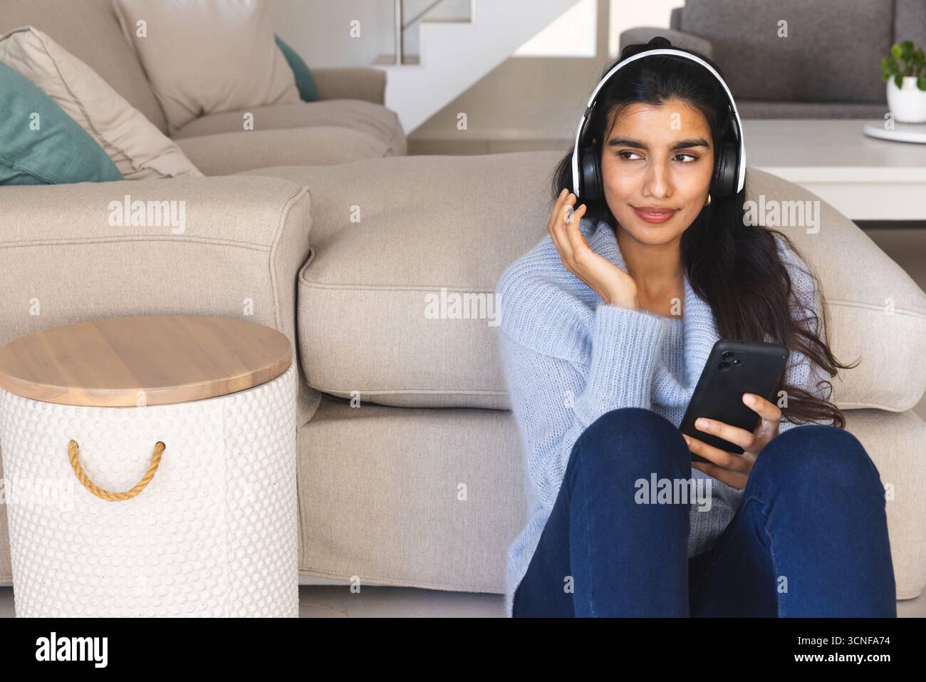 Südasiatische Frau hört zu Hause mit Kopfhörern auf dem Smartphone und sitzt vor dem Kopierraum des Sofas Stockfoto