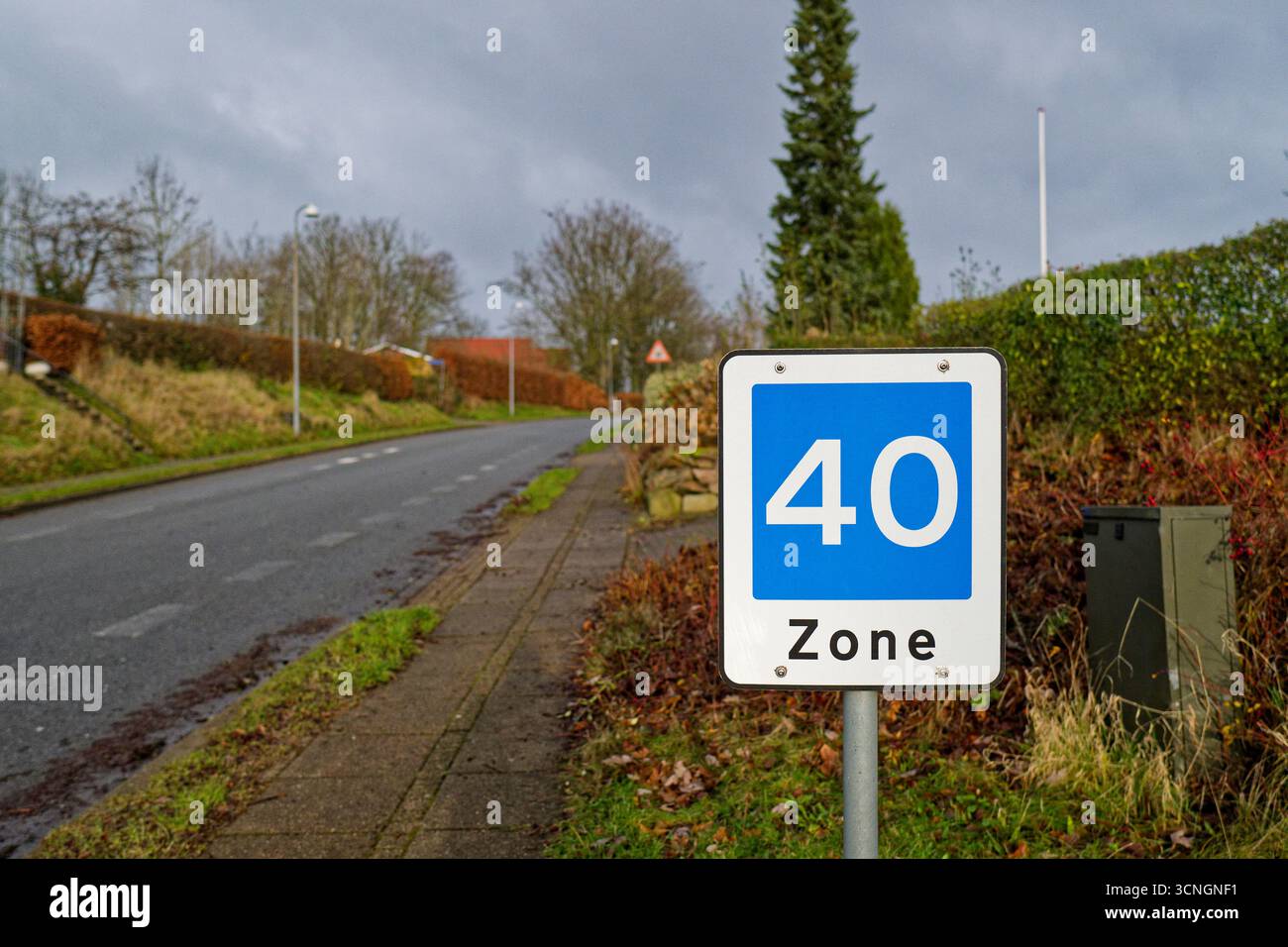 Ein Schild für die Zone 40 steht neben einer kleinen Trafohütte in der Nähe eines Bürgersteigs. Die Straße führt durch eine ruhige dänische Straße, feuchter Asphalt und kurzes Gras umrahmen den Stockfoto