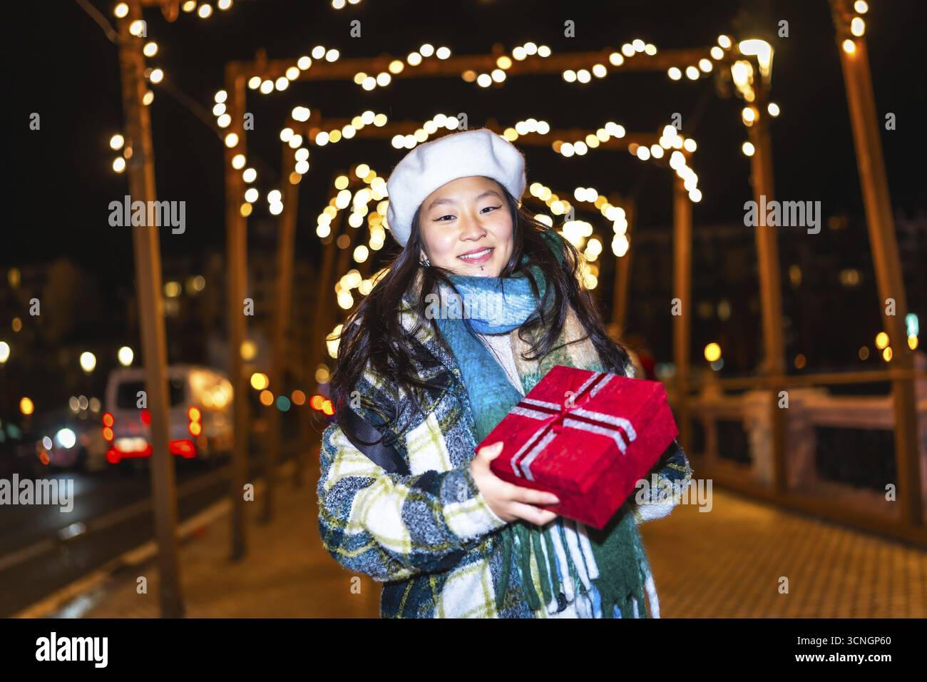 Lächelnde junge Frau, die weihnachtsgeschenk hält und die Winterferien unter dekorativen Lichtern in der Stadtstraße bei Nacht genießt Stockfoto