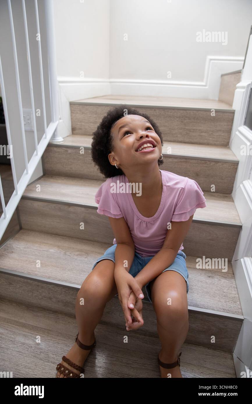 Afroamerikanisches Mädchen, das auf hölzernen Treppen im Haus sitzt, lächelnd neben Metall-Handgeländern Stockfoto