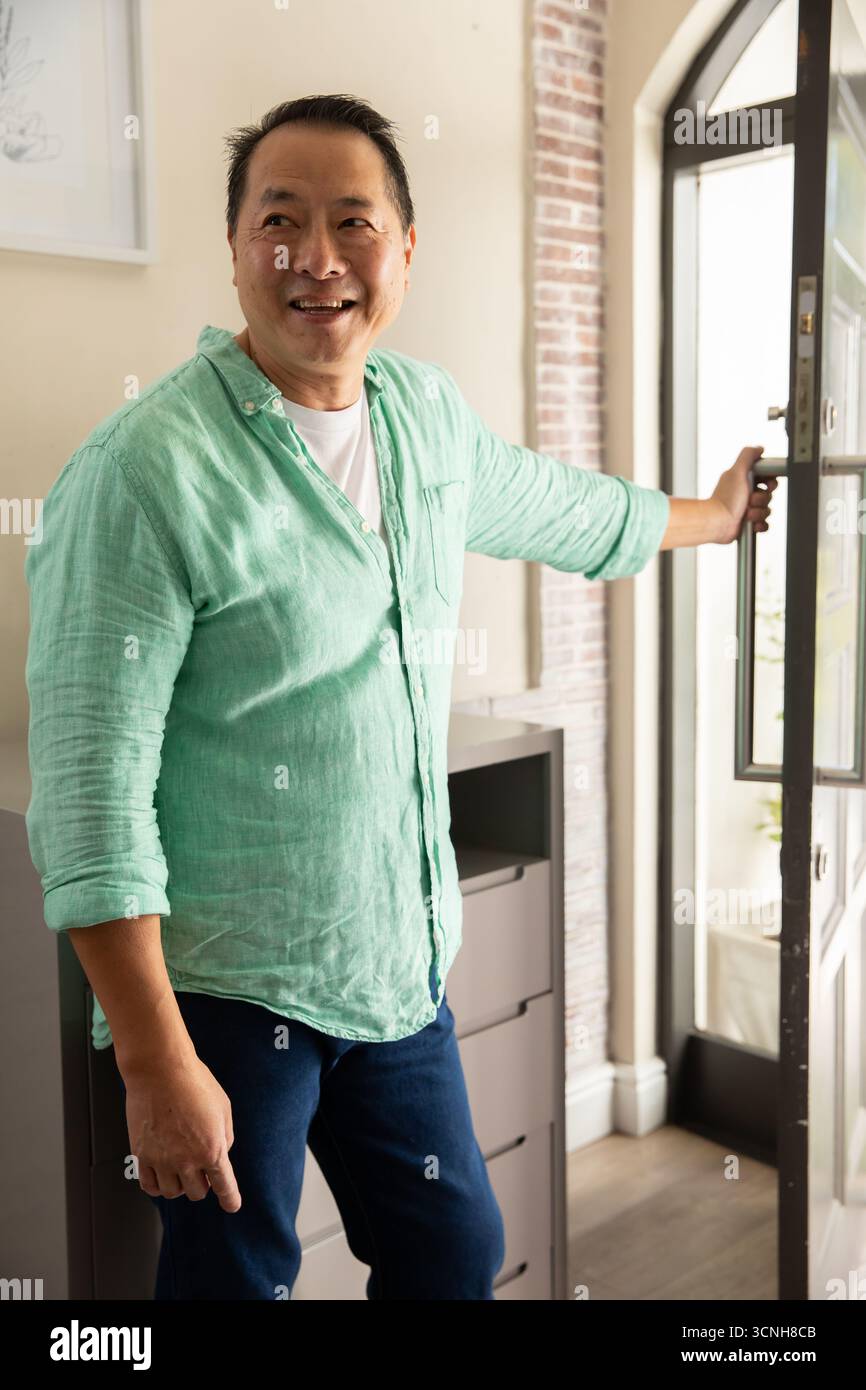 Asiatischer Mann, der im Eingangsbereich steht und eine Glastür neben der Kommode mit Lampe hält Stockfoto