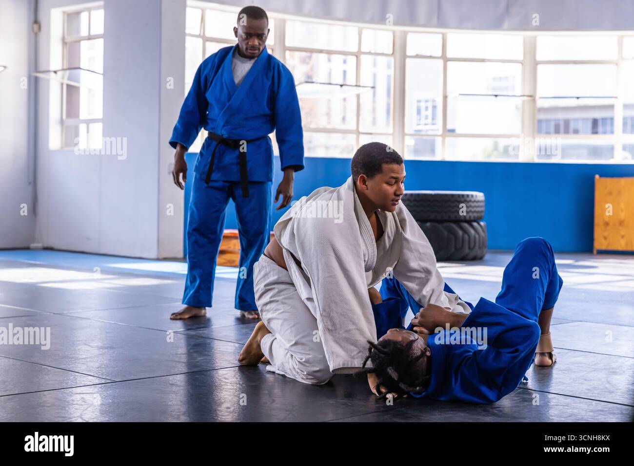 Verschiedene männliche Kampfsportlehrer, die das Grappling im gis mit zwei Schülern auf der Matte betreuen Stockfoto