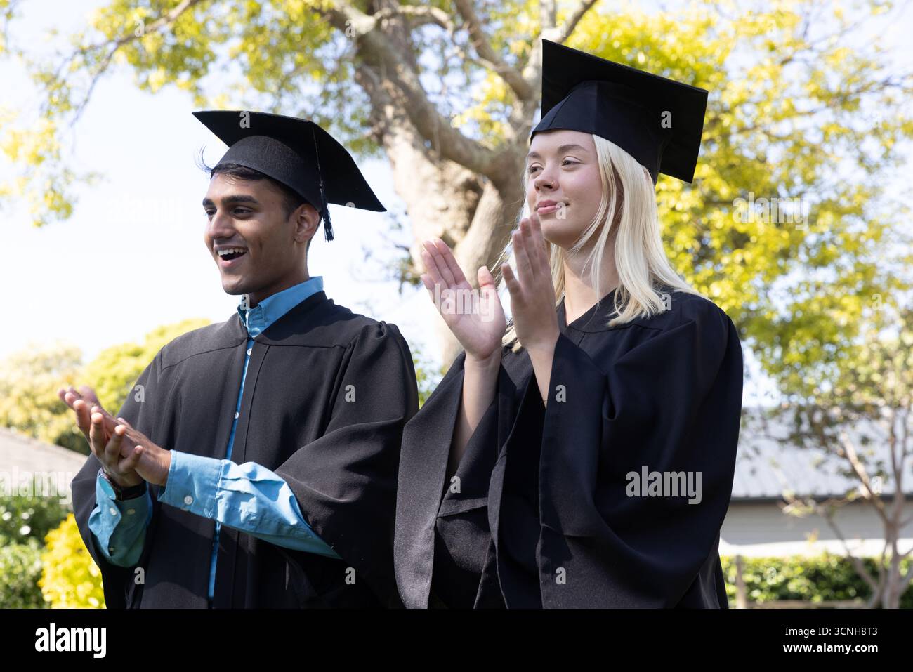 Verschiedene Absolventen in schwarzen Kleidern applaudieren den Beginn auf dem Campus-Rasen, Mörtelmützen Stockfoto