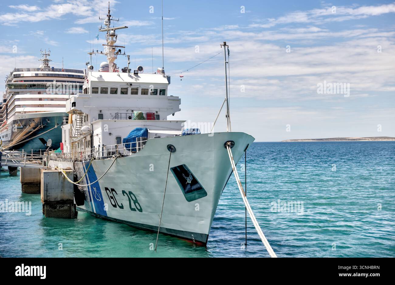 Ein Schiff der argentinischen Küstenwache legte neben einem Kreuzfahrtschiff am Puerto Madryn Pier an, mit türkisfarbenem Wasser und patagonischer Küste im Hintergrund Stockfoto