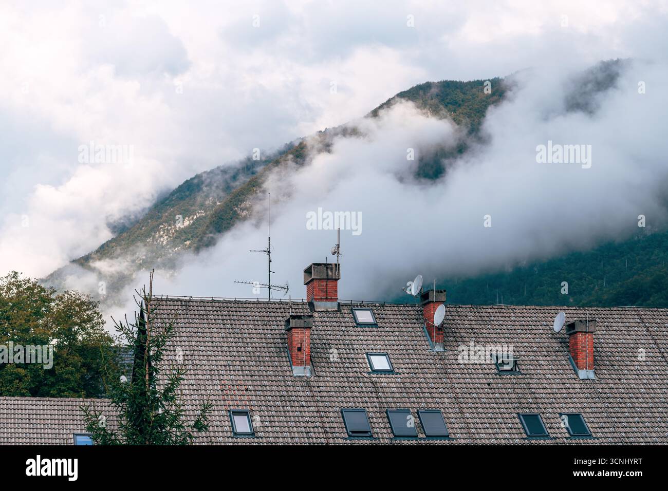 Dachplatte aus Zementfliesen mit Kamin und Dachfenster in einem Gebäude in der slowenischen Alpenlandschaft, mit Nebel, der sanft über die Umgebung steigt Stockfoto