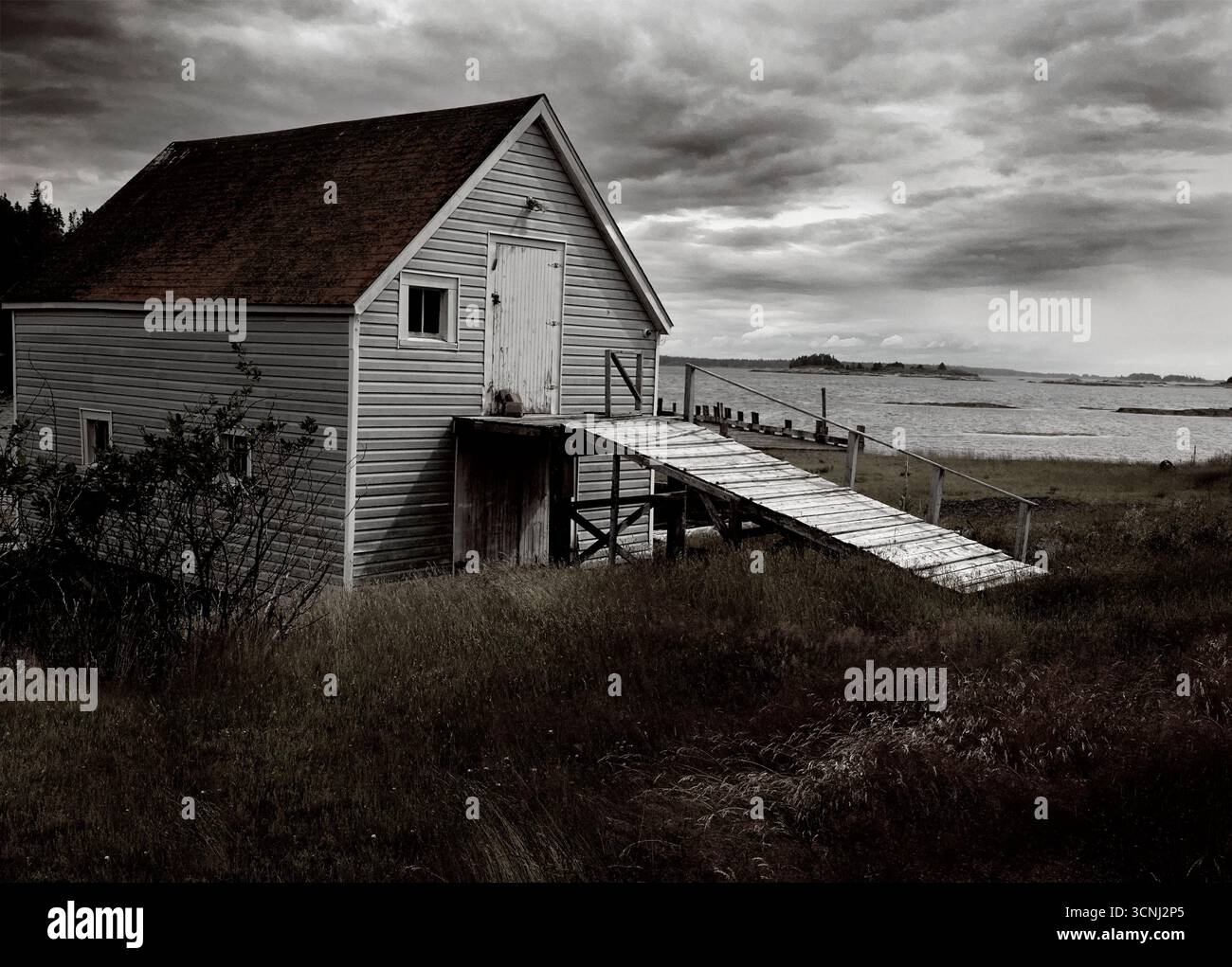Ein rustikaler Bootsschuppen liegt in der Nähe einer ruhigen Wasserstraße von Nova Scotia, umgeben von hohem Gras und bewölktem Himmel. Die Struktur verfügt über eine einfache Rampe, die zu führt Stockfoto