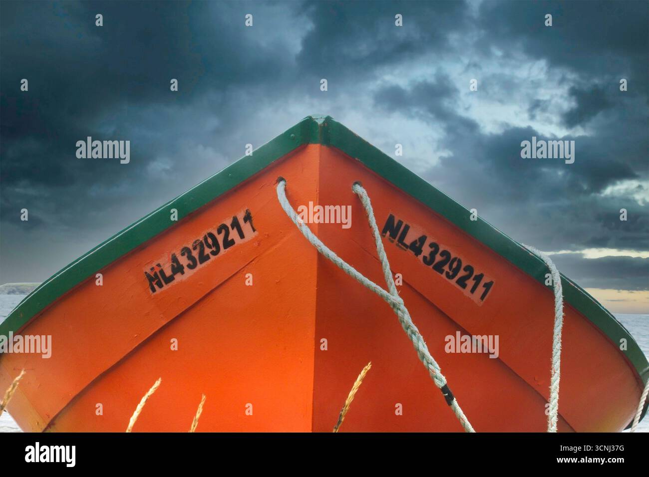 Ein dramatisches Bild eines orangen Bootes mit grüner Verkleidung sitzt ungenutzt am Sandstrand. Dunkle Wolken erheben sich über dem Himmel, während die Dämmerung den Himmel färbt und einen Drall erzeugt Stockfoto