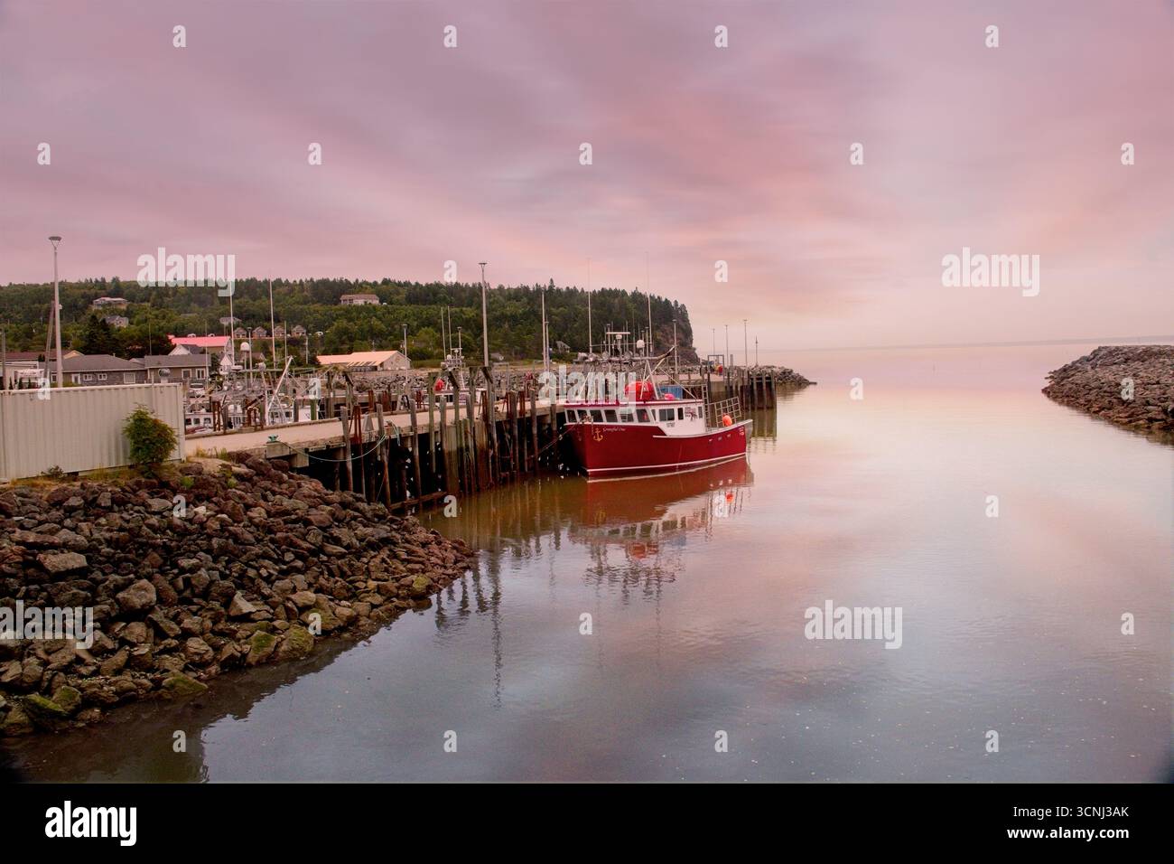 Ein leuchtend rotes Fischerboot liegt bei Flut in Alma, New Brunswick, Kanada, bei Sonnenaufgang sicher an. Der pastellfarbene Himmel schafft eine friedliche Atmosphäre Stockfoto