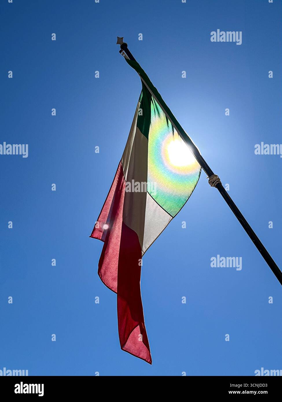 Italienische Flagge auf dem Pfosten, hinterleuchtet von der Sonne vor einem hellblauen Himmel Stockfoto