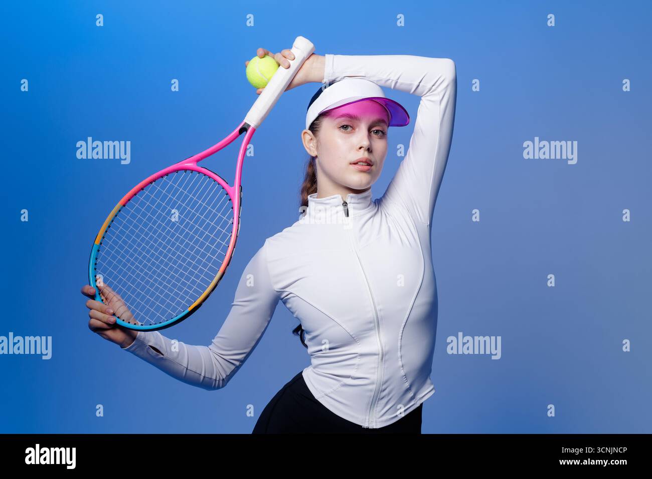 Selbstbewusste Tennisspielerin posiert mit Schläger und Ball im Studio auf blauem Hintergrund, bereit für Training und Wettkampf Stockfoto