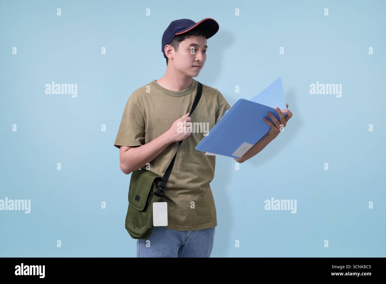 Ein lässiger junger Mann in einer Mütze hält blaue Mappen und steht im Profil mit einer grünen Tasche. Perfekt für Bildung, Studentenleben und alltägliche jugendleben Stockfoto