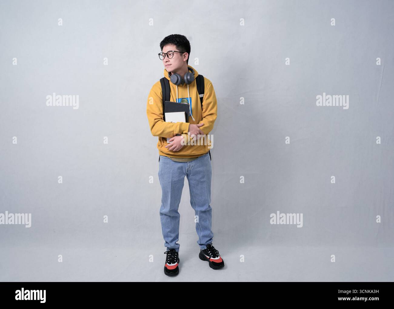 Ein selbstbewusster Schüler in einem Senfpullover steht mit Rucksack, Kopfhörern und Notebooks vor einem neutralen Hintergrund Stockfoto