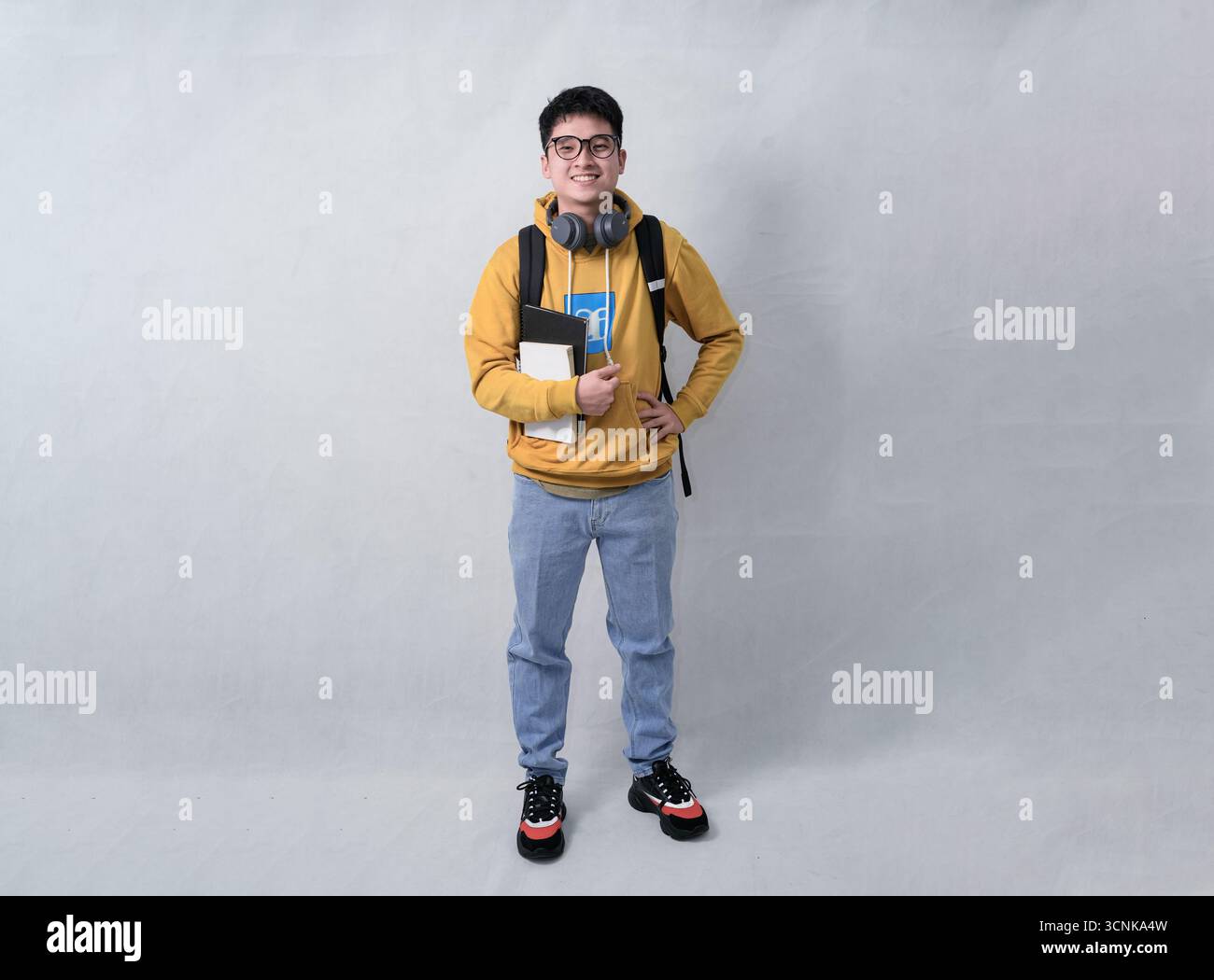 Ein selbstbewusster Schüler in einem Senfpullover steht mit Rucksack, Kopfhörern und Notebooks vor einem neutralen Hintergrund Stockfoto
