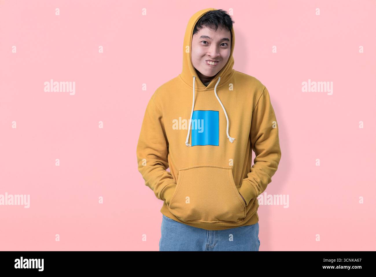 Ein fröhlicher junger Mann, der einen Senf-Hoodie mit einer blauen quadratischen Grafik trägt, steht vor einem sanften rosa Hintergrund Stockfoto