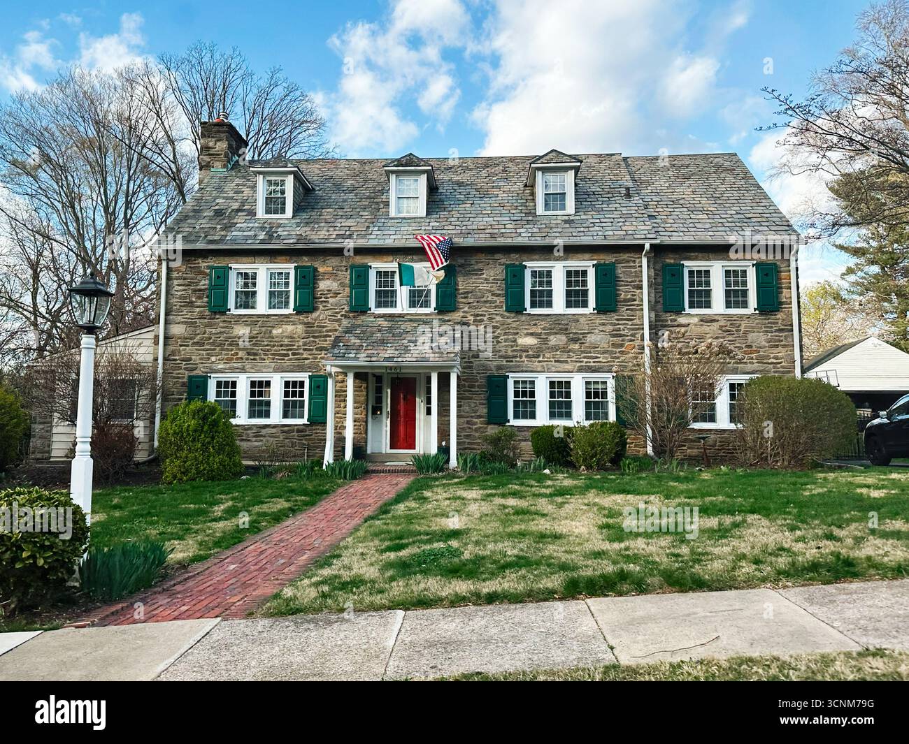 Philadelphia, Pennsylvania, USA - 28. April 2025: Zweistöckiges Steinhaus mit grünen Fensterläden, Backsteinpfaden und amerikanischer Flagge im sonnigen Vorhof. Stockfoto