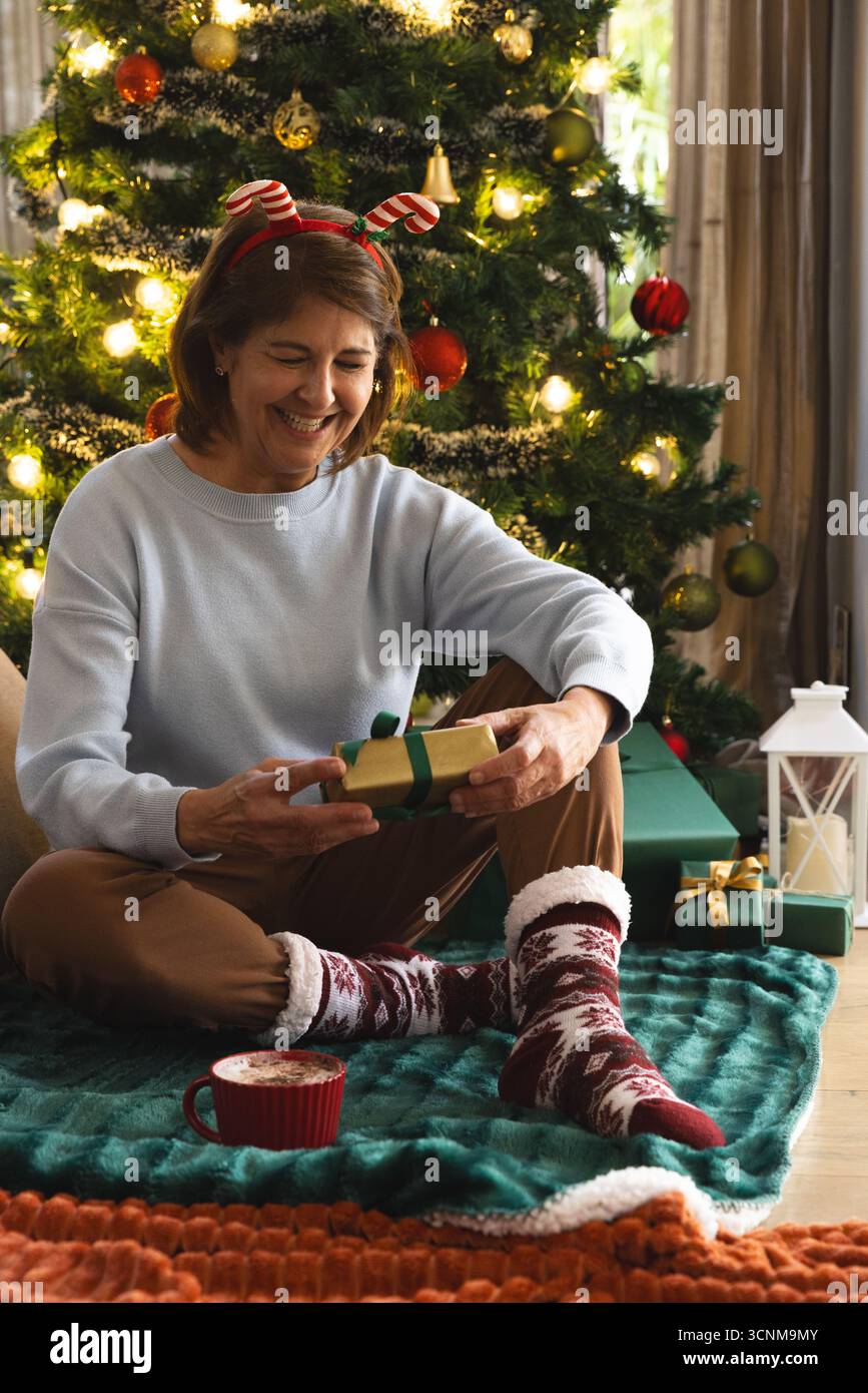 Hispanische Frau mit Geweih-Stirnband und Öffnung Gold gewickeltes Geschenk am Weihnachtsbaum auf Decke Stockfoto