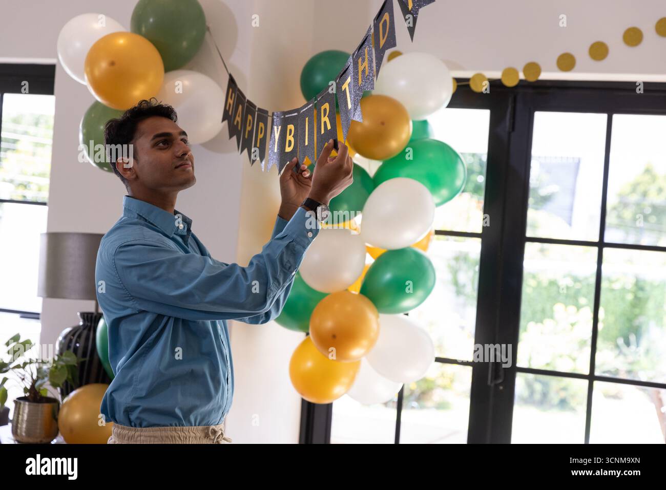 Indischer Mann, der Happy Birthday Banner im Wohnzimmer in der Nähe von Ballons hängt Stockfoto