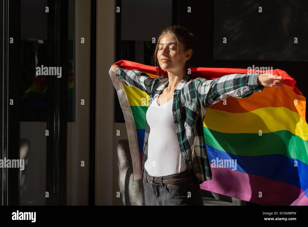 Frau, die im Wohnbereich bei Glasplatten steht, mit Regenbogenfahne hinter dem Rücken unter Sonnenlicht Stockfoto