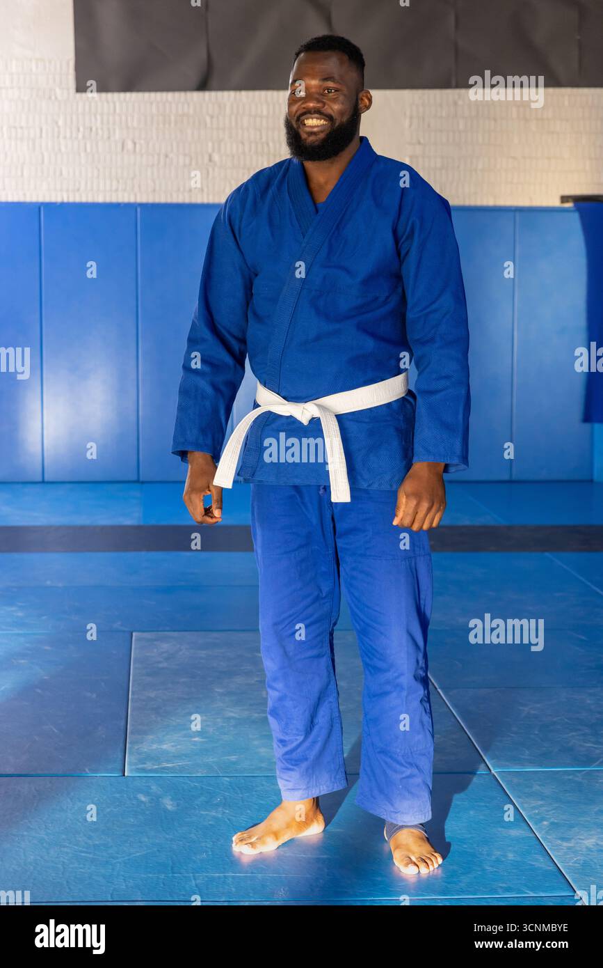 Lächelnder afroamerikaner, der auf Matten im Trainingsraum steht, in blauem Gi mit weißem Gürtel Stockfoto